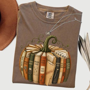 May include: A brown crew neck t-shirt featuring a graphic of a pumpkin constructed from stacked books with titles like True Crime, Missing Clues, and Case Files. The design includes a green stem and a thin silver chain necklace resting above it.