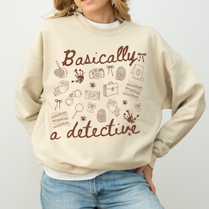 Könnte beinhalten: Beiges Sweatshirt mit dem Schriftzug "Basically a detective" in brauner Schrift. Das Design umfasst Detektiv-Illustrationen wie Fingerabdrücke, eine Lupe und Handschellen.