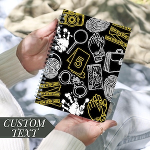 May include: A spiral-bound notebook with a black cover featuring crime scene illustrations in white and yellow. The notebook includes a camera, magnifying glass, fingerprints, handcuffs, and the text "CRIME SCENE DO NOT CROSS". The words "CUSTOM TEXT" are also visible.