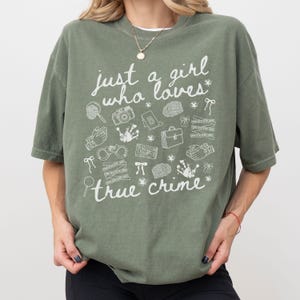 以下が含まれることがあります： 「just a girl who loves true crime」の白文字がプリントされたオリーブグリーンのTシャツ。虫眼鏡、カメラ、手錠など、犯罪に関連するイラストが描かれています。カジュアルで快適なスタイル。