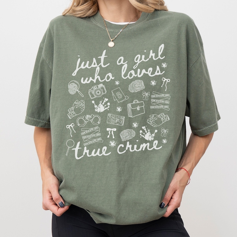 以下が含まれることがあります： 「just a girl who loves true crime」の白文字がプリントされたオリーブグリーンのTシャツ。虫眼鏡、カメラ、手錠など、犯罪に関連するイラストが描かれています。カジュアルで快適なスタイル。