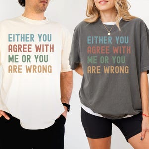 May include: Two short-sleeved t-shirts, one cream and one charcoal grey, each displaying the text "EITHER YOU AGREE WITH ME OR YOU ARE WRONG" in a stacked layout. The text is in teal, rust, green, and tan.