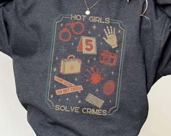 May include: Dark gray sweatshirt with a graphic that reads "HOT GIRLS SOLVE CRIMES." The graphic includes handcuffs, a handprint, a camera, a suitcase, a fingerprint, and the number 5 in a badge-like design.