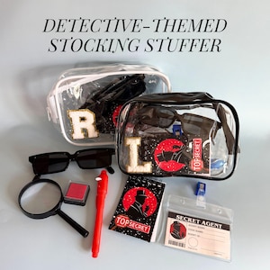 May include: Detective-themed stocking stuffer set with clear bags, sunglasses, magnifying glass, red ink pad, pen, notepad, and a secret agent ID. The bags have the letters "R" and "L". The notepad says "Top Secret".