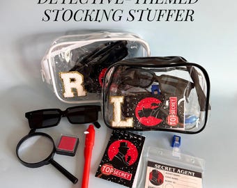 May include: Detective-themed stocking stuffer set with clear bags, sunglasses, magnifying glass, red ink pad, pen, notepad, and a secret agent ID. The bags have the letters "R" and "L". The notepad says "Top Secret".