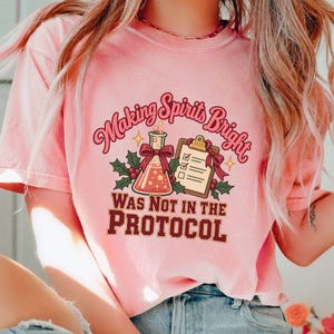 May include: A light pink t-shirt featuring a graphic with a laboratory flask, a clipboard, and holly leaves. The text reads "Making Spirits Bright WAS NOT IN THE PROTOCOL" in a mix of pink and dark red lettering with decorative stars.