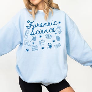Könnte beinhalten: Hellblaues Sweatshirt mit dem Schriftzug "Forensic Science" in Schreibschrift. Das Sweatshirt zeigt verschiedene blaue grafische Illustrationen, darunter Handschellen, eine Lupe, eine Kamera und einen Fingerabdruck.