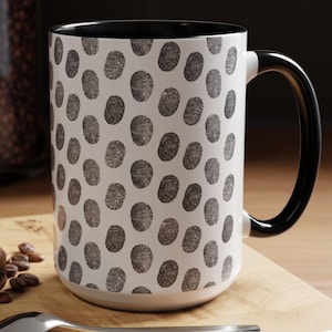 Puede incluir: Una taza de café de cerámica blanca con interior y asa negros. La taza está cubierta con un patrón de huellas dactilares negras. La taza está sobre una superficie de madera.