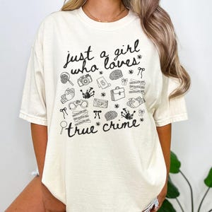 以下が含まれることがあります： 「just a girl who loves true crime」の黒い文字がプリントされたクリーム色のTシャツ。虫眼鏡、手錠、指紋など、犯罪解決に関連する様々な黒いイラストが描かれています。