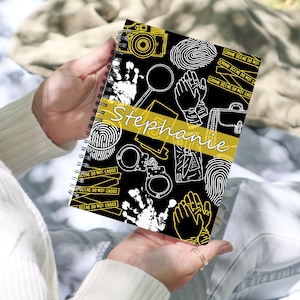 May include: A spiral-bound notebook with a black cover featuring white and yellow crime scene illustrations. The notebook includes a camera, magnifying glass, handcuffs, fingerprints, and the name "Stephanie" in a yellow banner.