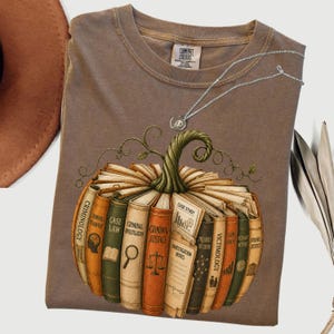 Könnte beinhalten: Ein braunes kurzärmeliges T-Shirt mit einer Grafik eines Kürbisses, der aus Büchern mit Titeln wie Criminology, Case Law und Criminal Justice besteht. Eine dünne silberne Halskette liegt am Halsausschnitt des T-Shirts.