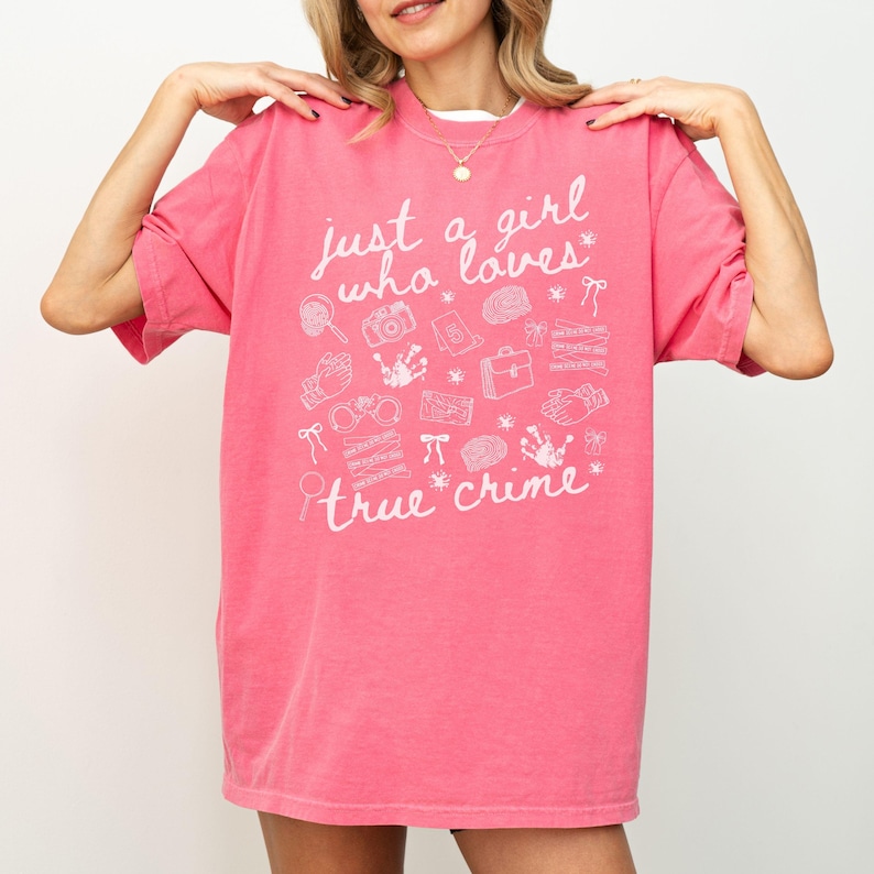 以下が含まれることがあります： 「just a girl who loves true crime」の白文字がプリントされたピンク色のTシャツ。手錠、虫眼鏡、カメラなど、犯罪捜査に関連する様々な白いグラフィックイラストが特徴です。