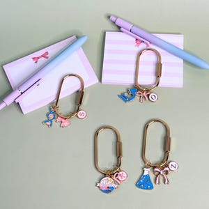 May include: Gold-colored keychains with science-themed charms, including a microscope, DNA strand, and a flask. Each keychain has a different letter charm and a pink bow. The background includes pink striped notepads and pens.