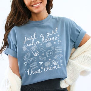 以下が含まれることがあります： 「just a girl who loves true crime」の白い文字が入った青いTシャツ。虫眼鏡、カメラ、指紋など、犯罪に関連する様々なイラストが描かれています。袖は折り返されています。