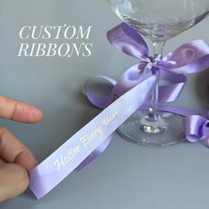 May include: A clear wine glass adorned with a light purple ribbon tied in a bow. The ribbon has the text "Hotter Every Year" printed on it. The words "Custom Ribbons" are in the background.