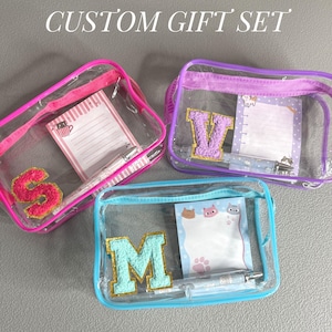 May include: Three clear plastic zippered pouches with colorful trim. Each pouch has a letter, S, V, and M, made of sparkly fabric inside. Each pouch also has a notepad and pen.