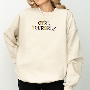 Può includere: Una felpa beige con la scritta ricamata "CTRL YOURSELF" in lettere colorate. La felpa ha un girocollo e maniche lunghe. Lo stile generale è casual e confortevole.