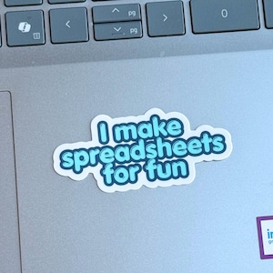 May include: A white sticker with the text "I make spreadsheets for fun" in a blue bubble font. The sticker is on a gray laptop, near the keyboard.