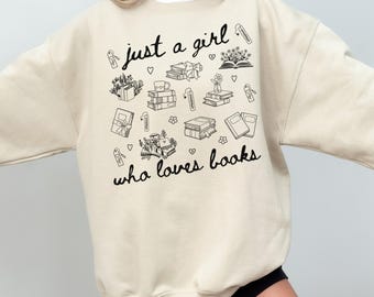 May include: Beige sweatshirt with the text "just a girl who loves books" in black script. The sweatshirt features black line drawings of books, gifts, flowers, and bookmarks. The overall design is simple and charming.