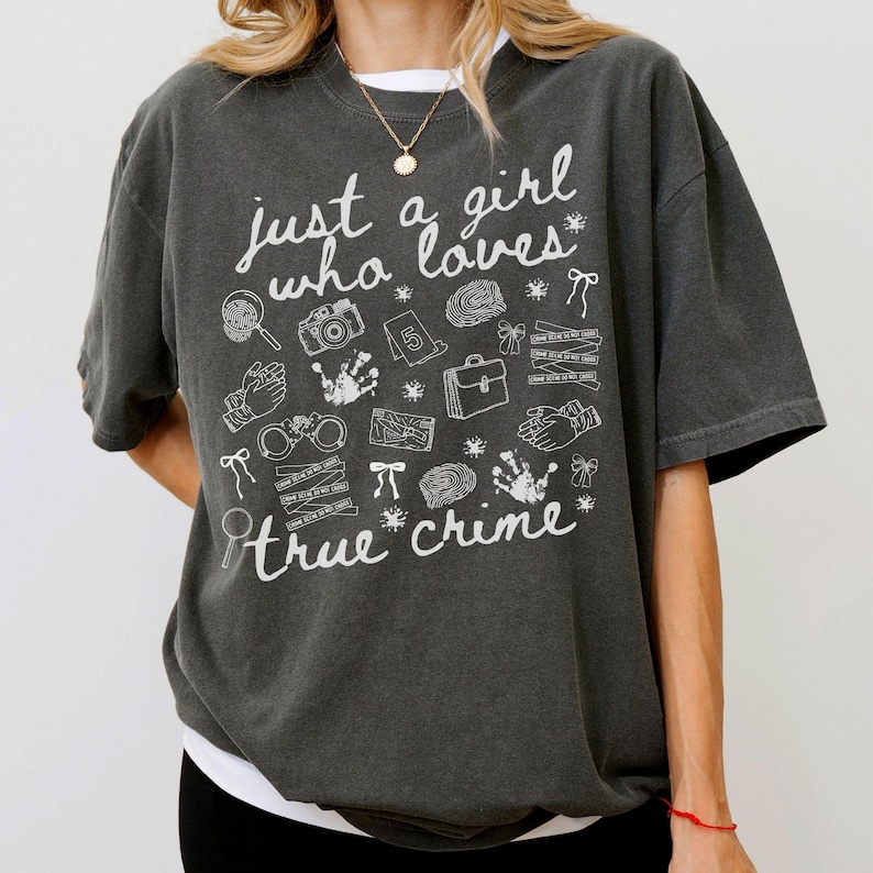 以下が含まれることがあります： 「just a girl who loves true crime」の白文字がプリントされた濃いグレーのTシャツ。虫眼鏡、カメラ、指紋、手錠など、犯罪に関連する様々なイラストが描かれています。