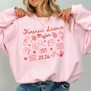 Könnte beinhalten: Hellrosa Sweatshirt mit rotem Aufdruck "Forensic Science Major", ergänzt durch rote Grafiken wie Handschellen, Lupe und Kamera. Das Jahr "2026" ist ebenfalls rot aufgedruckt.