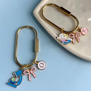 May include: Two gold-colored keychains with science-themed charms. One keychain has a microscope charm, a pink bow, and a white circle charm. The other keychain has a chemistry banner charm, a pink bow, and a pink circle charm.