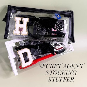 May include: Two clear zippered pouches, one black and one white, each containing items. The black pouch has sunglasses, a notebook, and a gold and white letter "H" patch. The white pouch has sunglasses, a red pen, and a gold and white letter "D" patch. Text reads "SECRET AGENT STOCKING STUFFER."