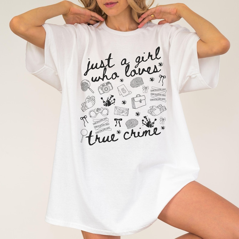 以下が含まれることがあります： 「just a girl who loves true crime」の文字と、虫眼鏡、カメラ、手錠など、犯罪に関連する様々な白黒イラストが描かれた白いTシャツ。