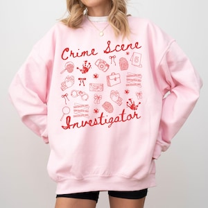 Könnte beinhalten: Hellrosa Sweatshirt mit dem Schriftzug "Crime Scene Investigator" in roter Schrift. Das Sweatshirt zeigt rote Illustrationen von forensischen Werkzeugen, darunter eine Lupe, eine Kamera und Handschellen. Das Gesamtdesign hat einen spielerischen, illustrativen Stil.