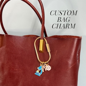 May include: A burgundy leather tote bag with a gold-colored carabiner clip and a charm. The charm features a blue microscope, a pink bow, and a pink circle. The text "CUSTOM BAG CHARM" is visible.