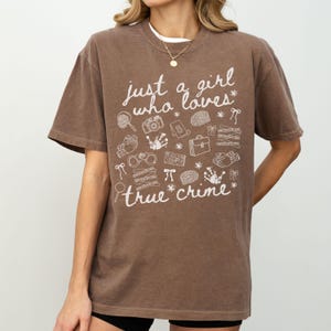 以下が含まれることがあります： 「just a girl who loves true crime」の白い文字がプリントされた茶色のTシャツ。虫眼鏡、カメラ、手錠など、犯罪に関連するイラストが描かれています。半袖でクルーネックです。