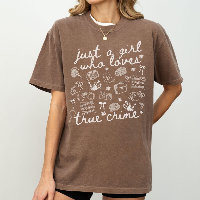 以下が含まれることがあります： 「just a girl who loves true crime」の白い文字がプリントされた茶色のTシャツ。虫眼鏡、カメラ、手錠など、犯罪に関連するイラストが描かれています。半袖でクルーネックです。