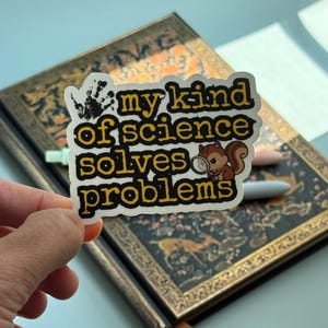 May include: A white sticker with the text "my kind of science solves problems" in yellow and black. A cartoon squirrel with a magnifying glass is on the right. A black handprint is on the left. The sticker is held over a decorative book.