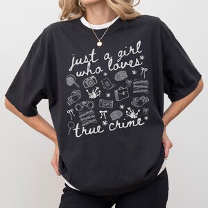 以下が含まれることがあります： 「just a girl who loves true crime」の白文字が入った黒いTシャツ。虫眼鏡、カメラ、手錠、指紋など、犯罪に関連するイラストが描かれています。