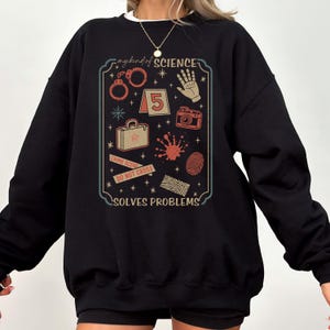 Könnte beinhalten: Schwarzes Sweatshirt mit einer Grafik mit Tatortelementen wie Handschellen, einem Fingerabdruck und einer Kamera. Der Text auf der Grafik lautet "my kind of SCIENCE SOLVES PROBLEMS". Das Design ist in Rot-, Beige- und Türkistönen gehalten.