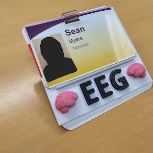 以下が含まれることがあります： 「Sean Myers」という名前と「Technician」という肩書きのIDバッジ。 バッジには写真があり、「EEG」という文字と2つのピンク色の脳の模型で飾られています。 黄色の蛍光ペンが背景に見えます。