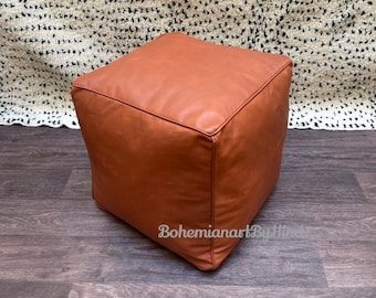 May include: A brown leather ottoman with a square shape. The ottoman is sitting on a wooden floor.