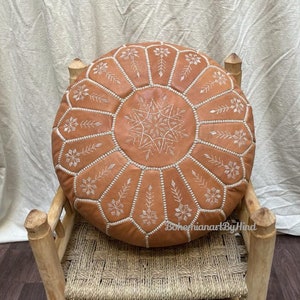 Puede incluir: Un puf redondo de cuero marr&oacute;n con bordados blancos en forma de flores. El puf est&aacute; sentado en una silla de madera con un asiento tejido.