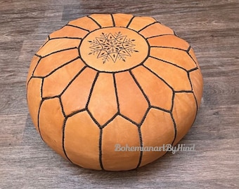 May include: A round, tan leather ottoman with a geometric design and black stitching. The top has a decorative starburst pattern. This floor-level seating option is suitable for a living room or bedroom.