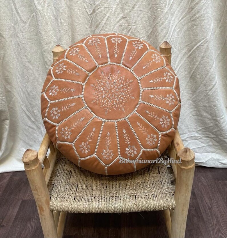 Puede incluir: Un puf redondo de cuero marr&oacute;n con bordados blancos en forma de flores. El puf est&aacute; sentado en una silla de madera con un asiento tejido.