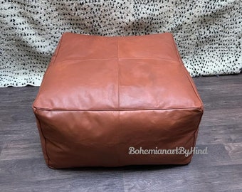May include: A brown leather pouf ottoman with a square shape and a stitched design. The pouf is sitting on a wooden floor. The text "BohemianartByHind" is visible on the pouf.