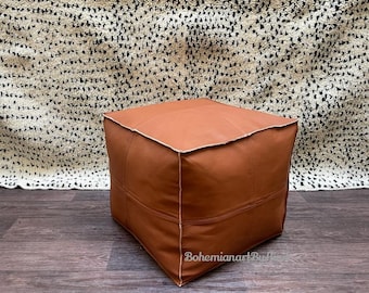 May include: A square brown leather ottoman with white stitching. The ottoman is sitting on a wooden floor.