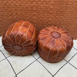 May include: Two brown leather poufs with intricate stitching and a star-shaped design in the center. The poufs are sitting on a white and black patterned rug.