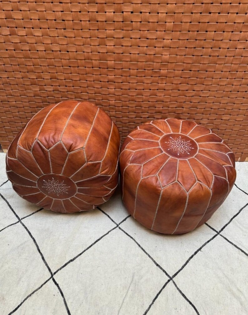 May include: Two brown leather poufs with intricate stitching and a star-shaped design in the center. The poufs are sitting on a white and black patterned rug.