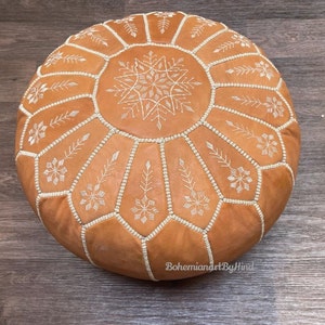 Puede incluir: Un puf de cuero marr&oacute;n con dise&ntilde;os de copo de nieve bordados en blanco. El puf es redondo y tiene una parte superior acolchada.