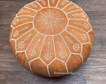 May include: A tan leather pouf with white embroidered snowflake designs. The pouf is round and has a tufted top.