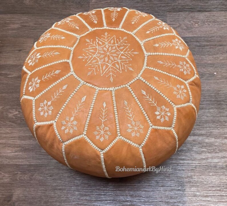 Puede incluir: Un puf de cuero marr&oacute;n con dise&ntilde;os de copo de nieve bordados en blanco. El puf es redondo y tiene una parte superior acolchada.