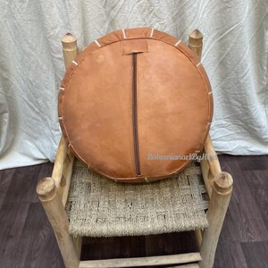 Puede incluir: Un puf redondo de cuero marr&oacute;n con costuras blancas se encuentra sobre una silla de madera con un asiento de paja trenzada. El puf tiene una cremallera en el lateral.