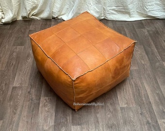 May include: A square, tan leather pouf ottoman featuring a grid-like panel construction with visible stitching along the edges. The surface displays a natural, smooth texture with subtle variations in color. The text BohemianartByHind is printed on the side.