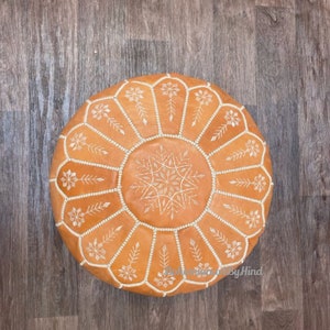 Puede incluir: Un puf redondo de cuero marr&oacute;n con dise&ntilde;os florales bordados en blanco. El puf est&aacute; sobre un suelo de madera.